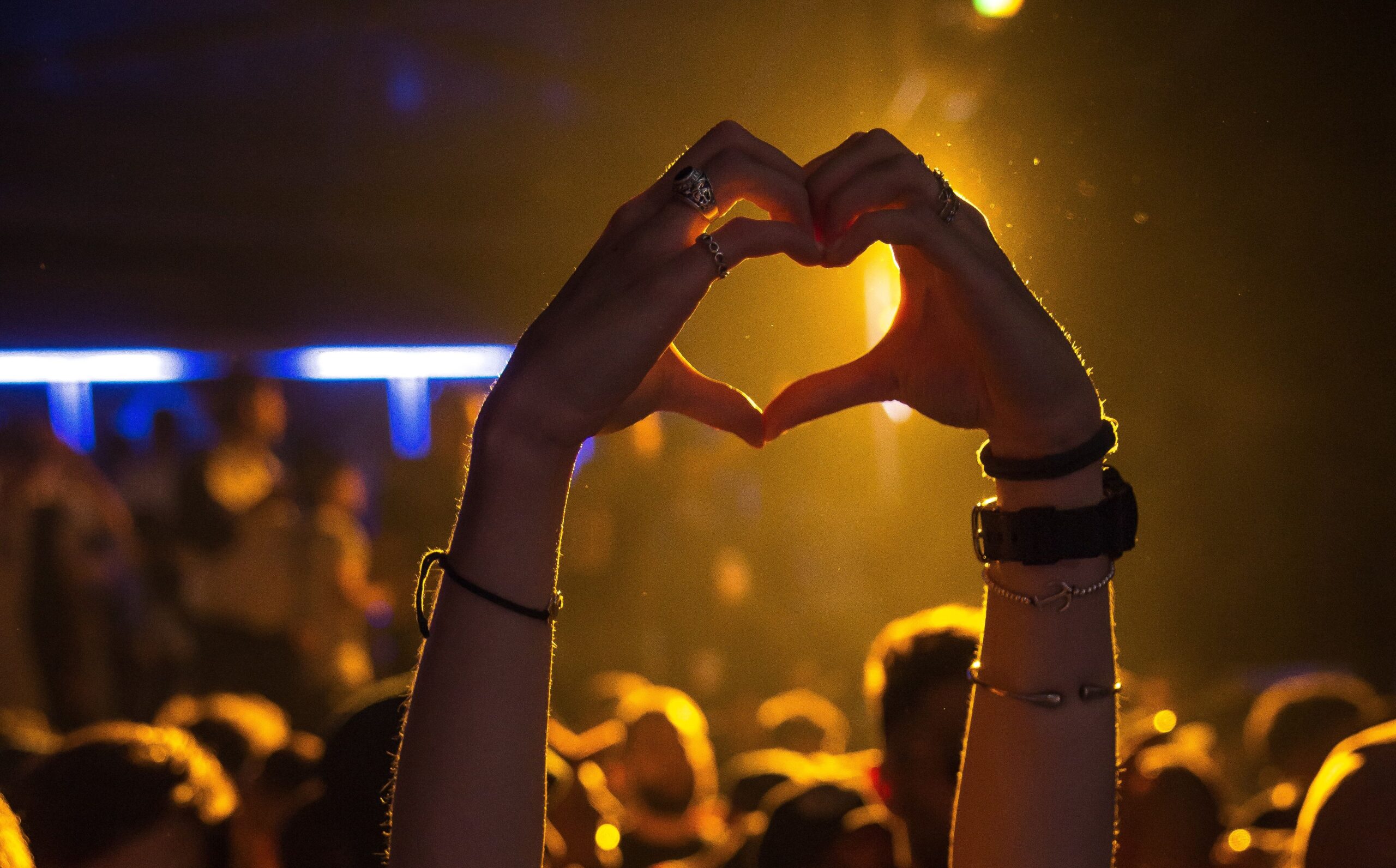 A person's hands form a heart shape against a backdrop of warm, golden concert lighting. The crowd below is softly blurred, with people illuminated by blue and yellow hues, creating an atmospheric and energetic scene. The focus is on the gesture, symbolizing love and connection to the music and the moment.