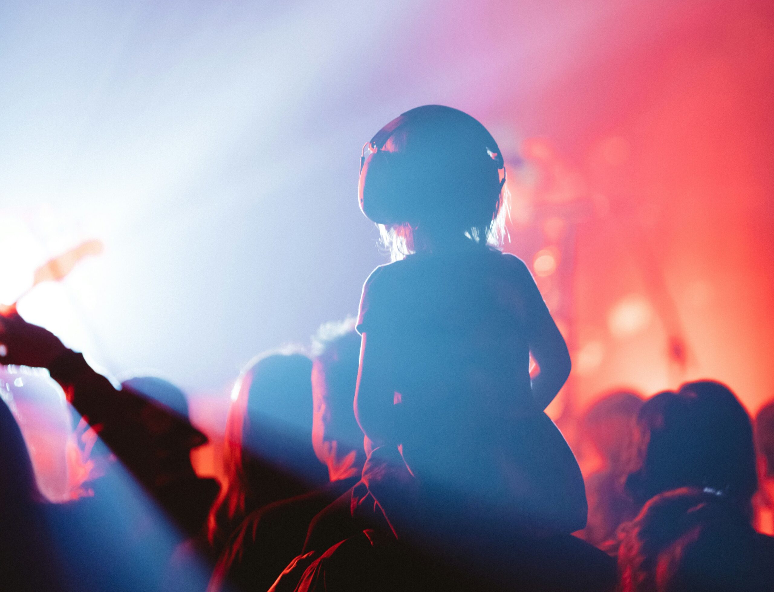 A child wearing large noise-canceling headphones sits on an adult's shoulders at a concert, surrounded by colorful stage lighting with vibrant blue and red beams cutting through the crowd.