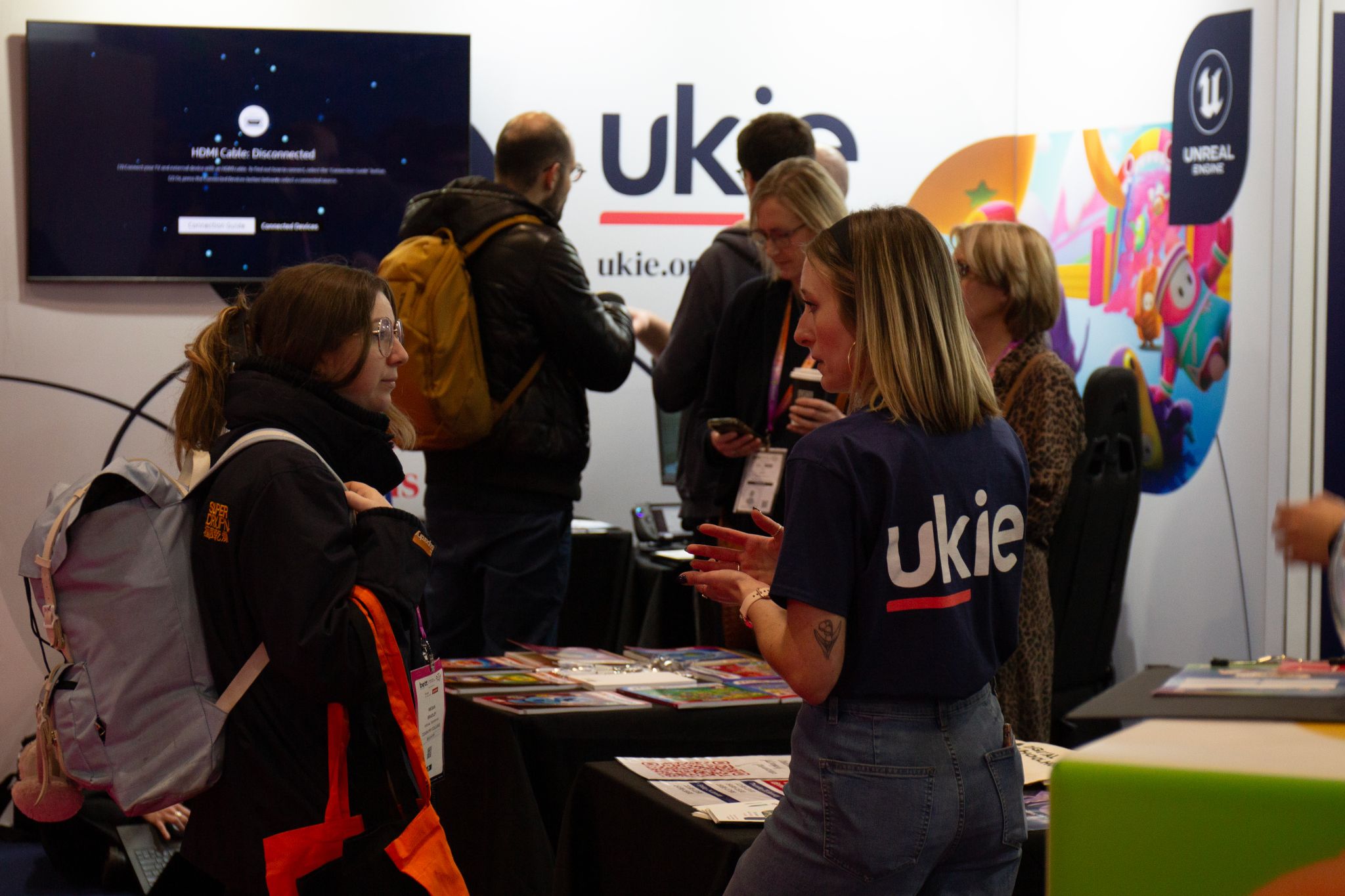 A bustling scene at an event booth hosted by Ukie, the UK's interactive entertainment industry body. A Ukie staff member, wearing a navy-blue shirt with the Ukie logo on the back, is engaged in conversation with an attendee carrying a backpack. In the background, several other attendees interact near displays and materials. The booth features vibrant signage, including a colorful Unreal Engine banner, and a screen displaying an HDMI error message. The atmosphere is lively, suggesting a networking or industry expo environment.