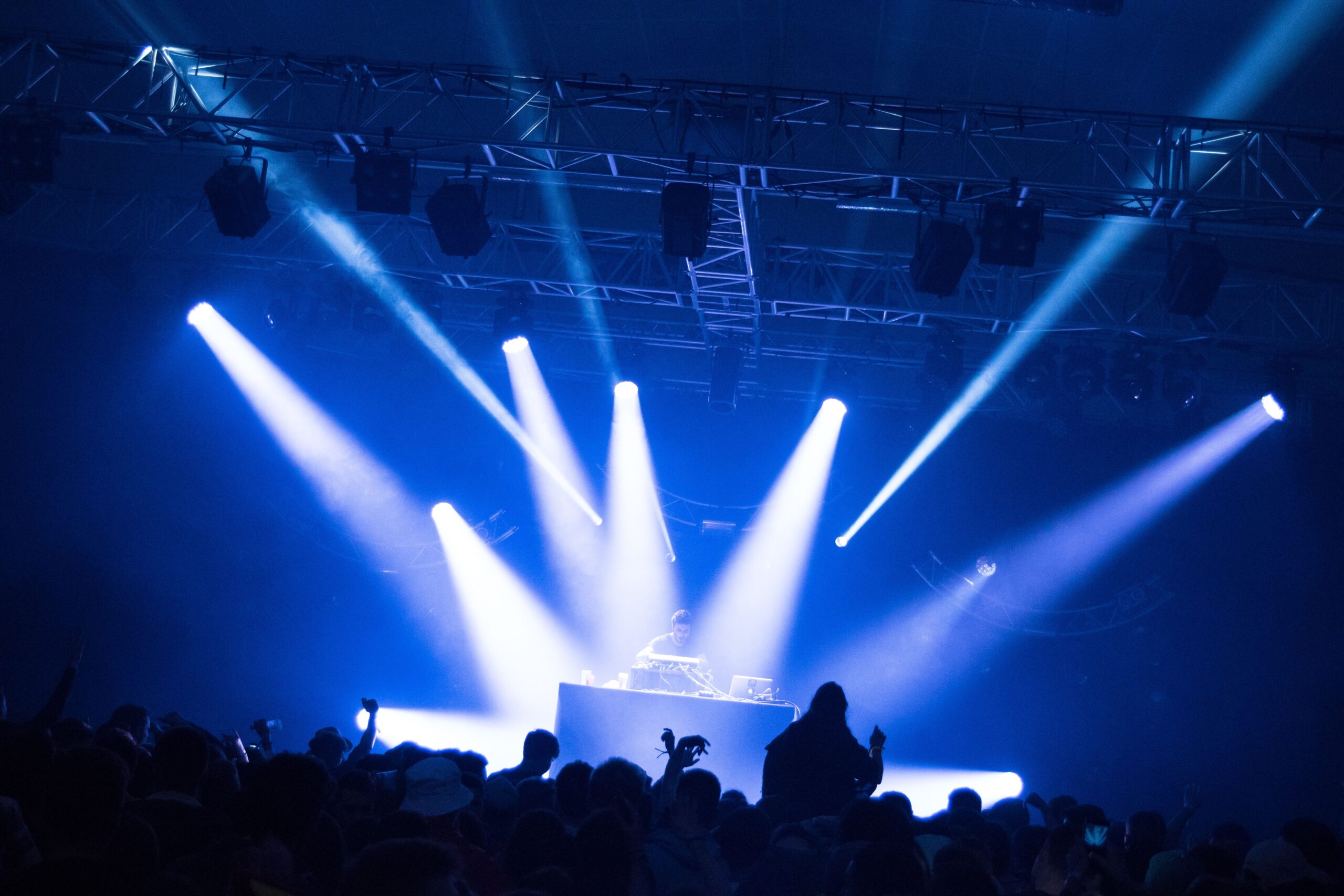 A DJ performs on stage under intense blue and white spotlight beams at a concert. The lights radiate from the ceiling, illuminating the DJ booth and casting a dramatic glow over the crowd below. Silhouetted audience members can be seen raising their hands and enjoying the energetic atmosphere. The scene highlights the vibrant lighting and immersive experience of a live music event.