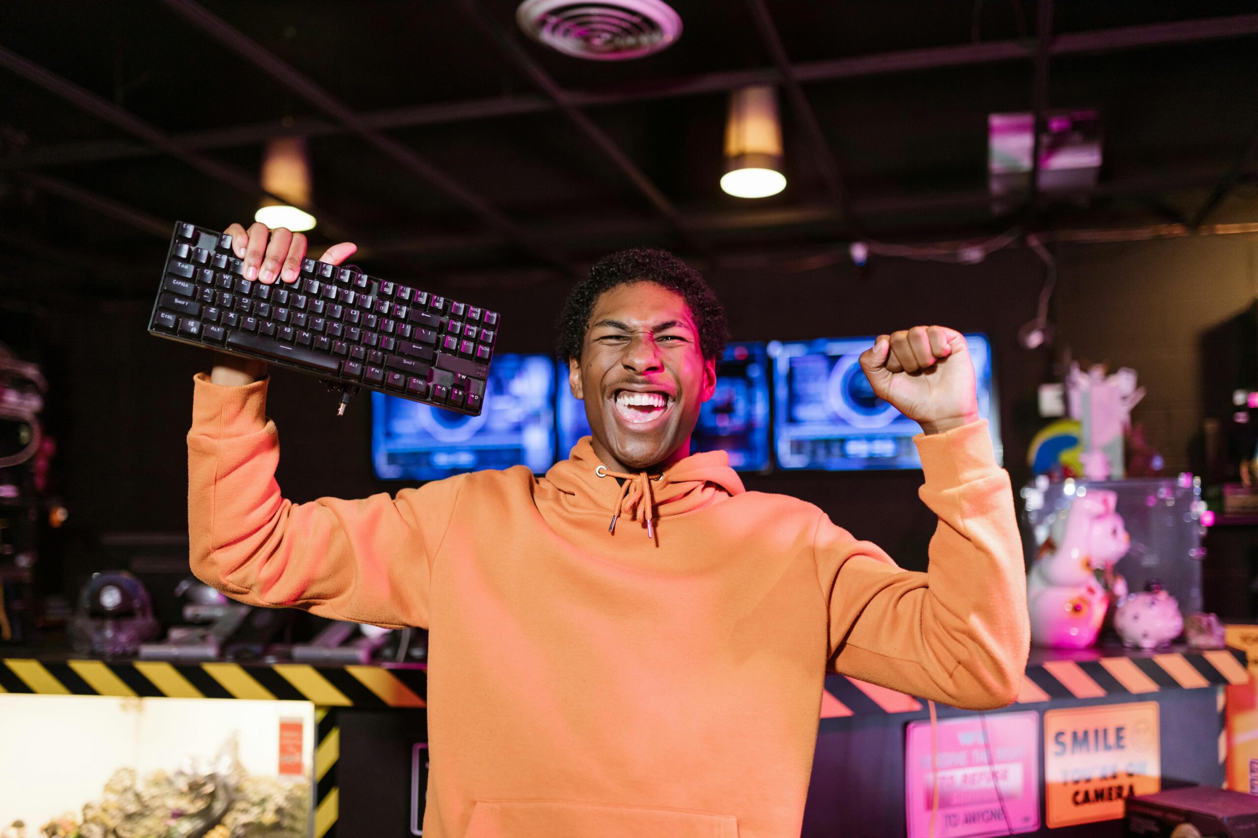 A young man in an orange hoodie celebrates enthusiastically, holding a keyboard aloft with one hand and clenching his other fist in excitement. He has a big smile on his face, expressing joy or victory. Behind him, a gaming setup with multiple screens and colorful equipment fills the room, contributing to a vibrant, high-energy atmosphere.