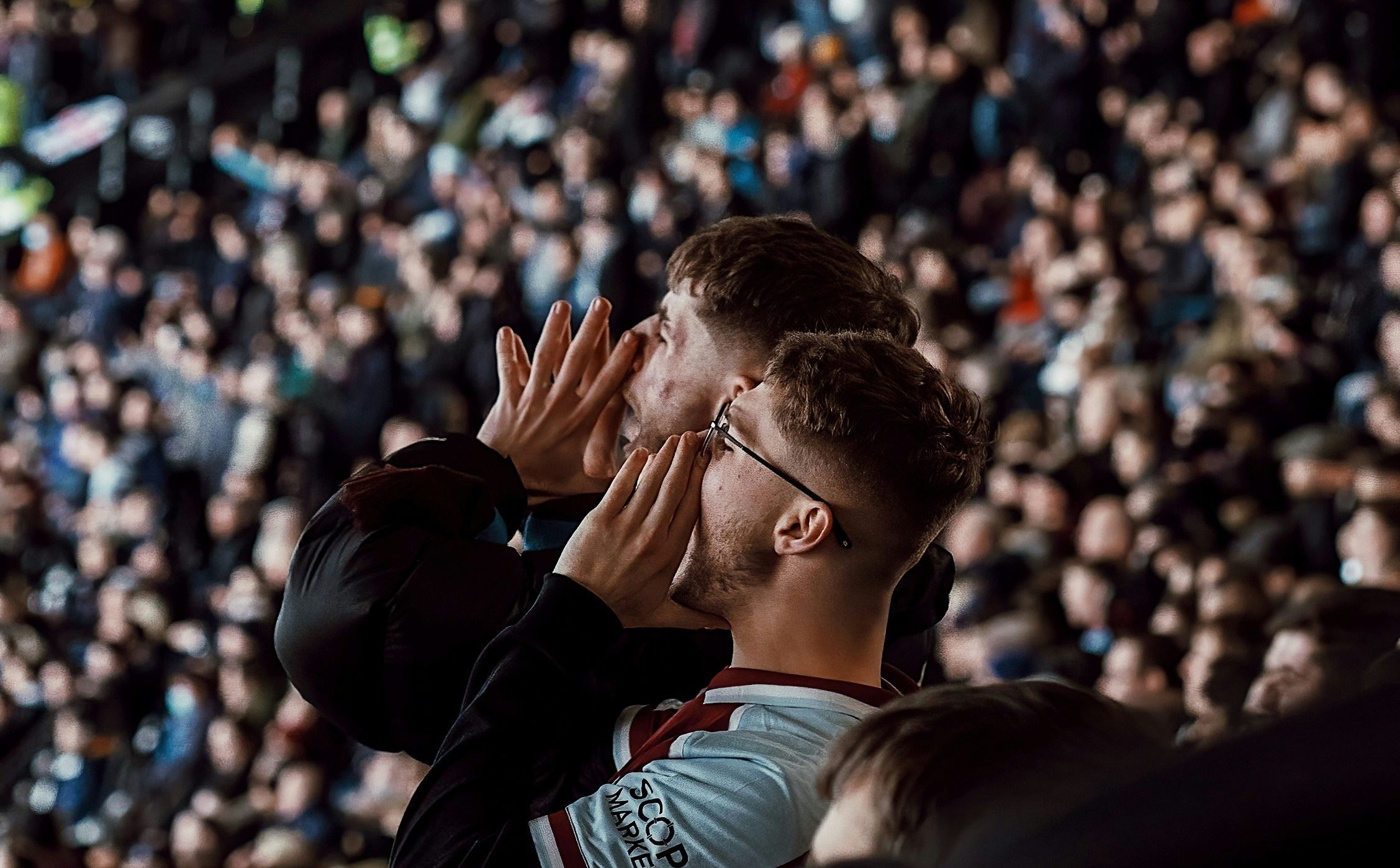 Two enthusiastic football fans shout and cheer from the stands of a crowded stadium. One fan wears a light blue and maroon football jersey, cupping his hands around his mouth as he yells. The background is filled with a blurred, lively crowd, capturing the intense atmosphere of a live sports event.