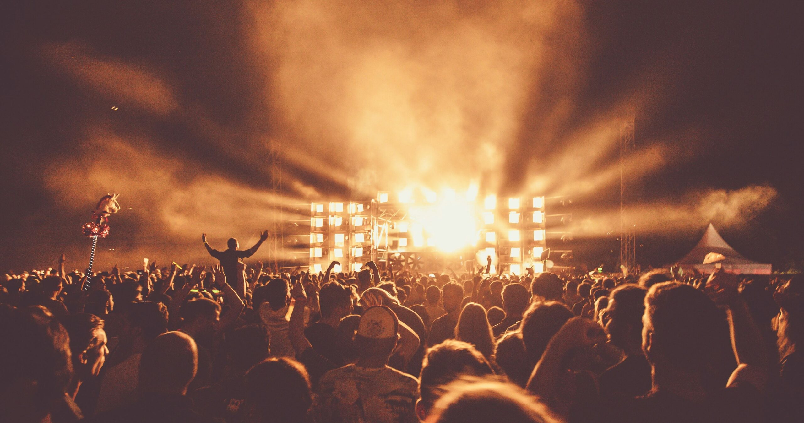 A large crowd of people gathered at a nighttime outdoor music festival, with bright, golden stage lights illuminating the scene. Concertgoers cheer with their hands raised, while thick smoke and atmospheric lighting create a dramatic effect. A whimsical unicorn decoration is raised above the crowd on a pole, adding a playful touch to the lively atmosphere.